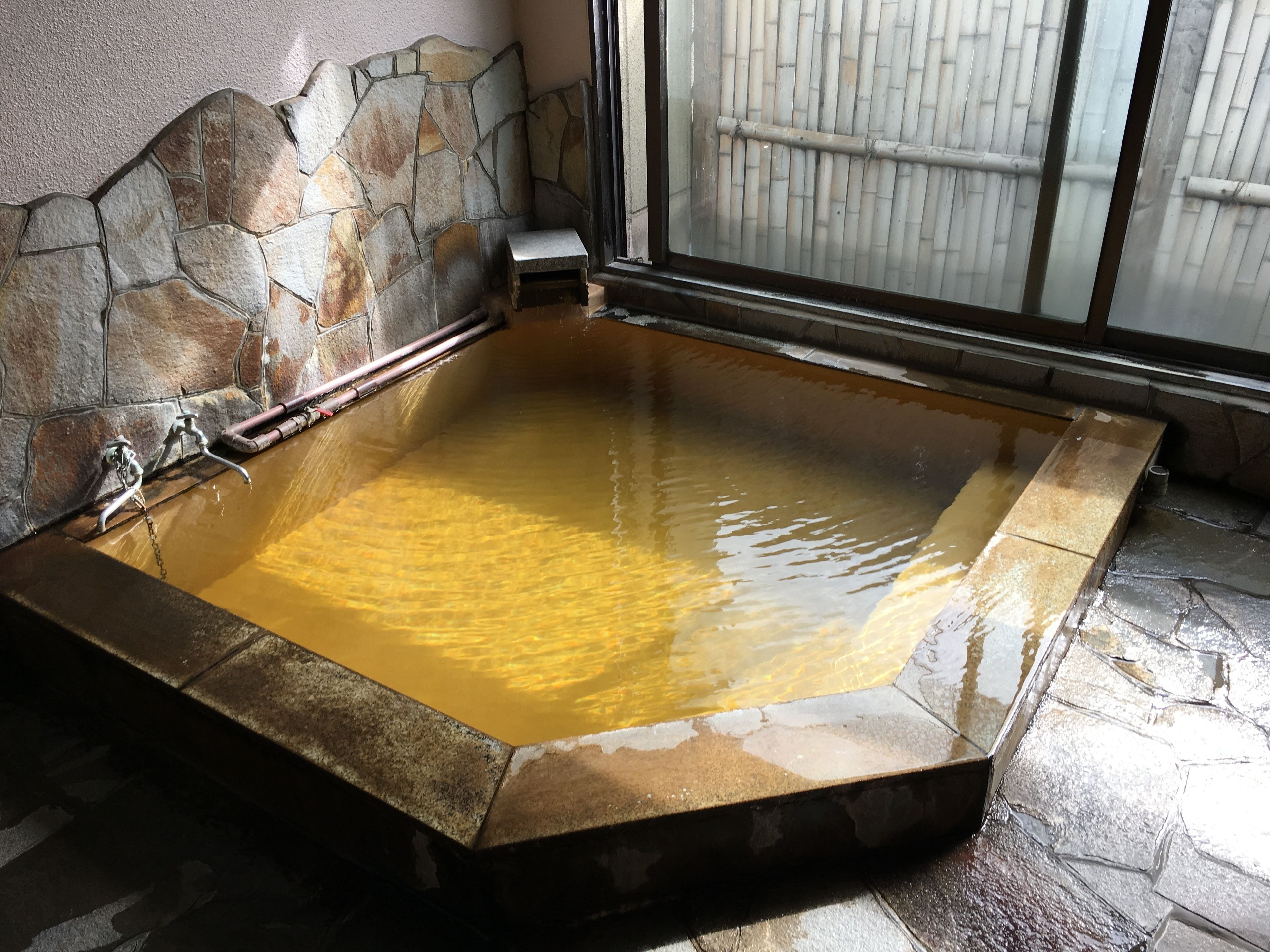 A stone-lined hot tub with a small wooden stool and a metal faucet.
