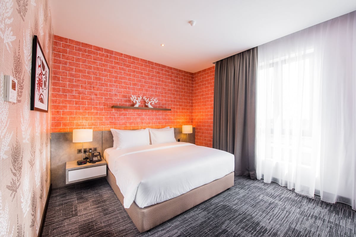 A hotel room with a brick wall, a large bed, two bedside tables with lamps, a nightstand, and a window with sheer curtains.