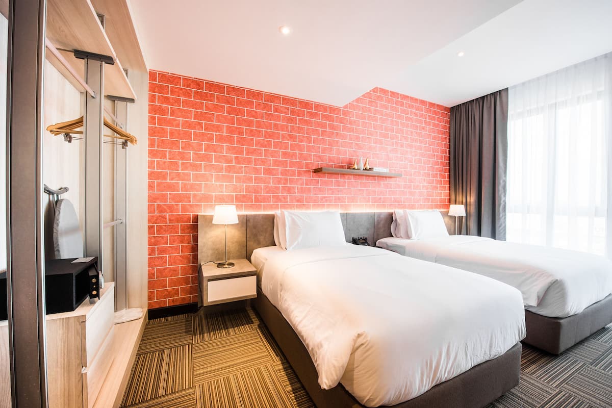 A hotel room with two beds, a red brick wall, a wooden floor, a mirror, and a shelf with decorative items.