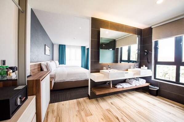 Una habitación de hotel moderna con una cama grande, un baño separado con un espejo amplio y suelo de madera.