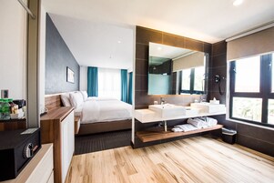 Una habitación de hotel moderna con una cama grande, un baño separado con un espejo amplio y suelo de madera.