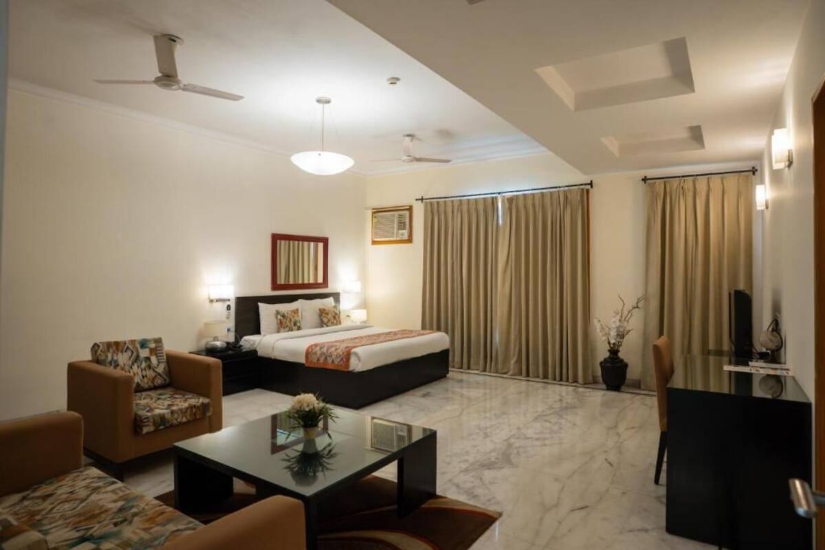 A hotel room with a bed, a desk, a chair, a coffee table, a ceiling fan, and a window with curtains.