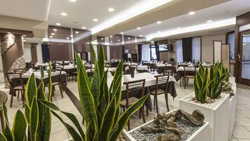 Ein geräumiges Restaurant mit mehreren Tischen, Stühlen, Topfpflanzen und einem Fernseher an der Wand.