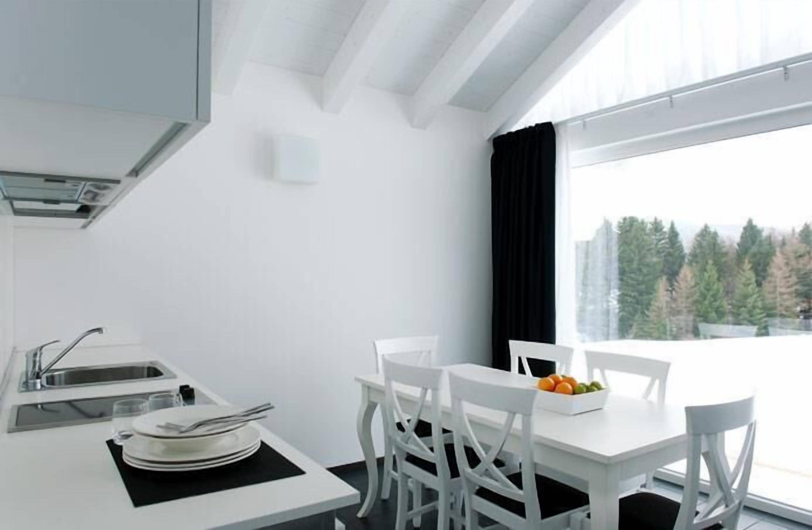 Una cocina moderna con armarios blancos, un comedor con sillas blancas y un amplio ventanal con vistas a árboles.