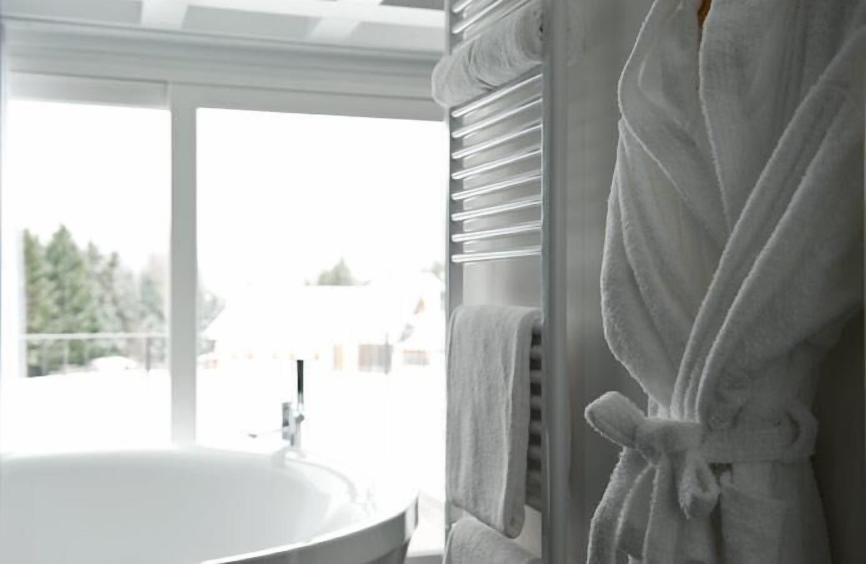 A bathroom with a white bathtub, a window with blinds, and a white bathrobe hanging on a rack.