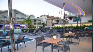 An outdoor seating area with tables and chairs, a pool, and a view of hotel buildings in the background. - Oasis Otel (Marmaris)