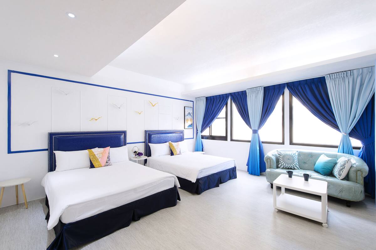 A hotel room with two beds, a sofa, a coffee table, and large windows with blue curtains.