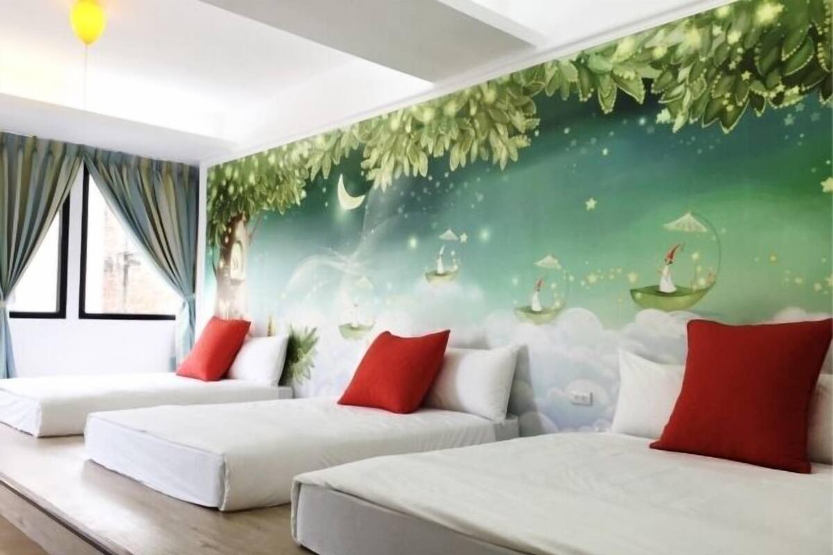 A hotel room with three beds, each with white bedding and red pillows. The room features a large mural with a nature theme, including trees and a moon.