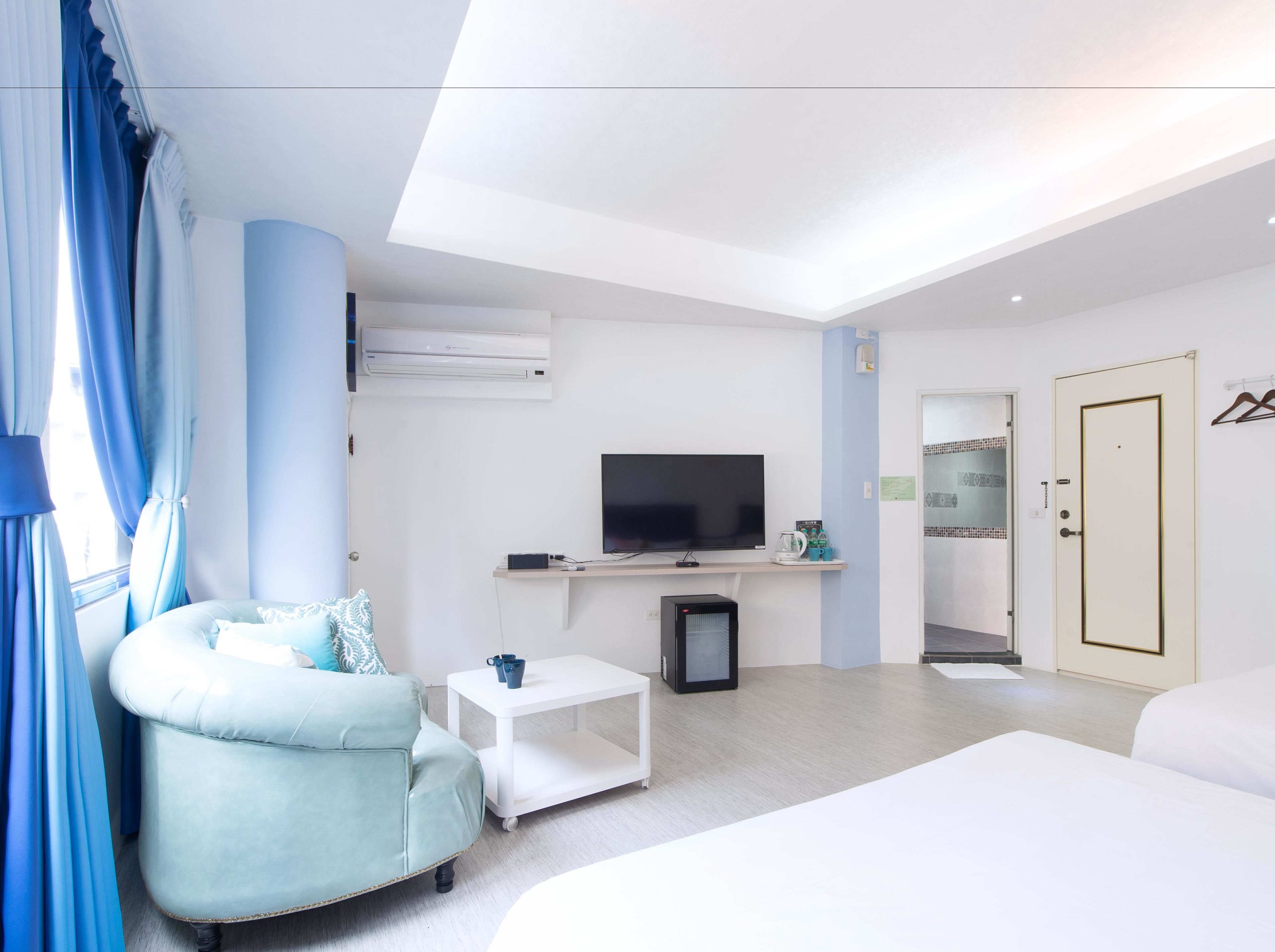 Una habitación de hotel moderna con una cama grande, un rincón de descanso con un sillón azul claro, una mesita blanca pequeña, un televisor de pantalla plana y un armario.