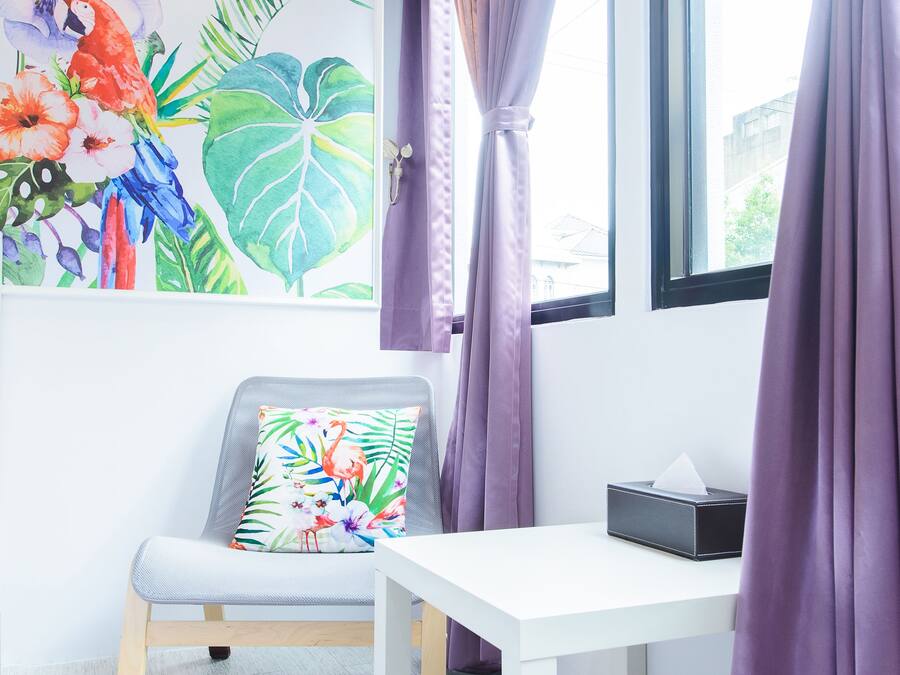 A room with a white table, a gray chair with a floral pillow, a window with purple curtains, and a wall with a colorful floral painting.