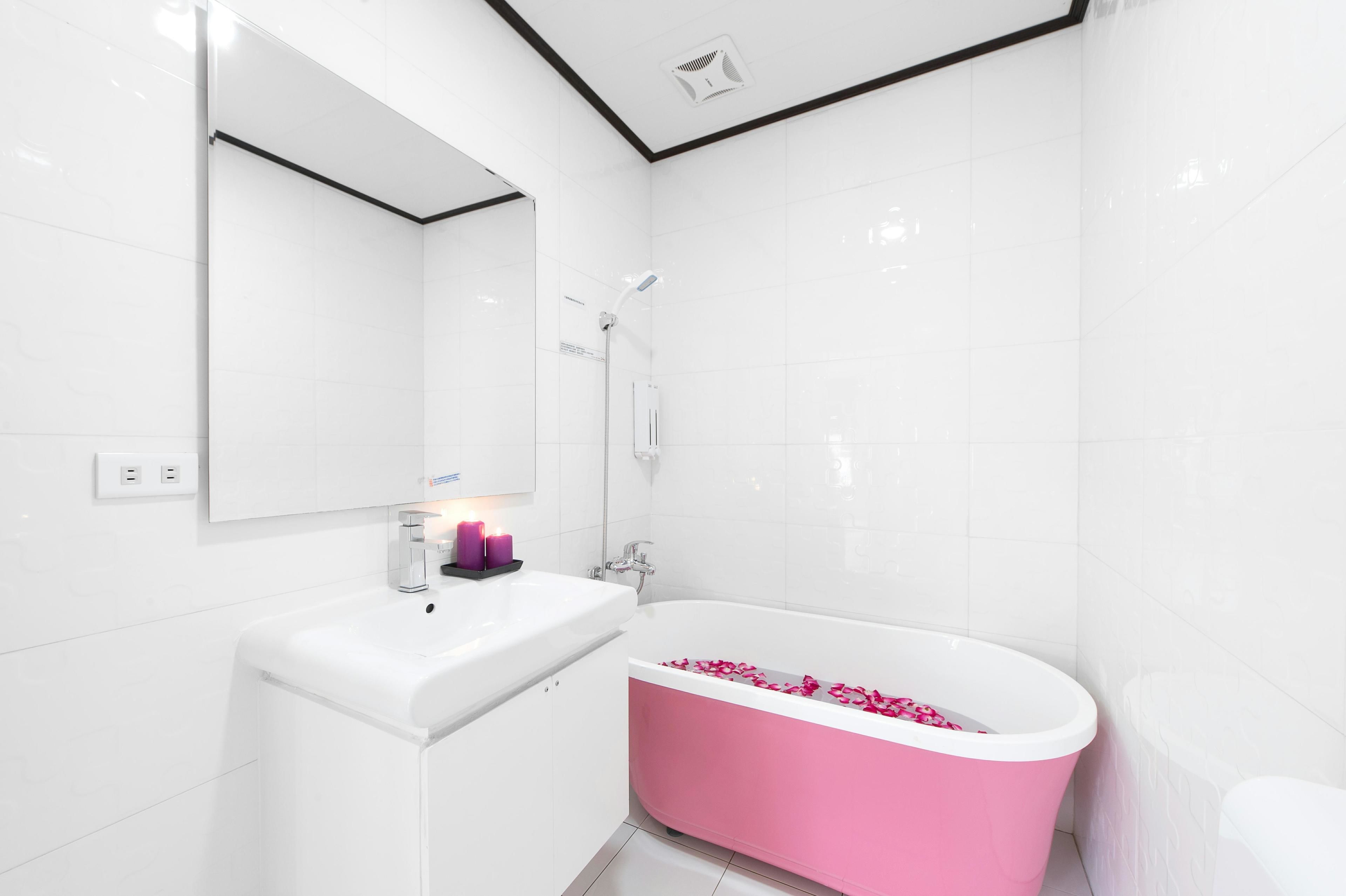 Un baño moderno con azulejos blancos, un lavamanos blanco, una bañera rosa y una ducha con cabezal de ducha manual.