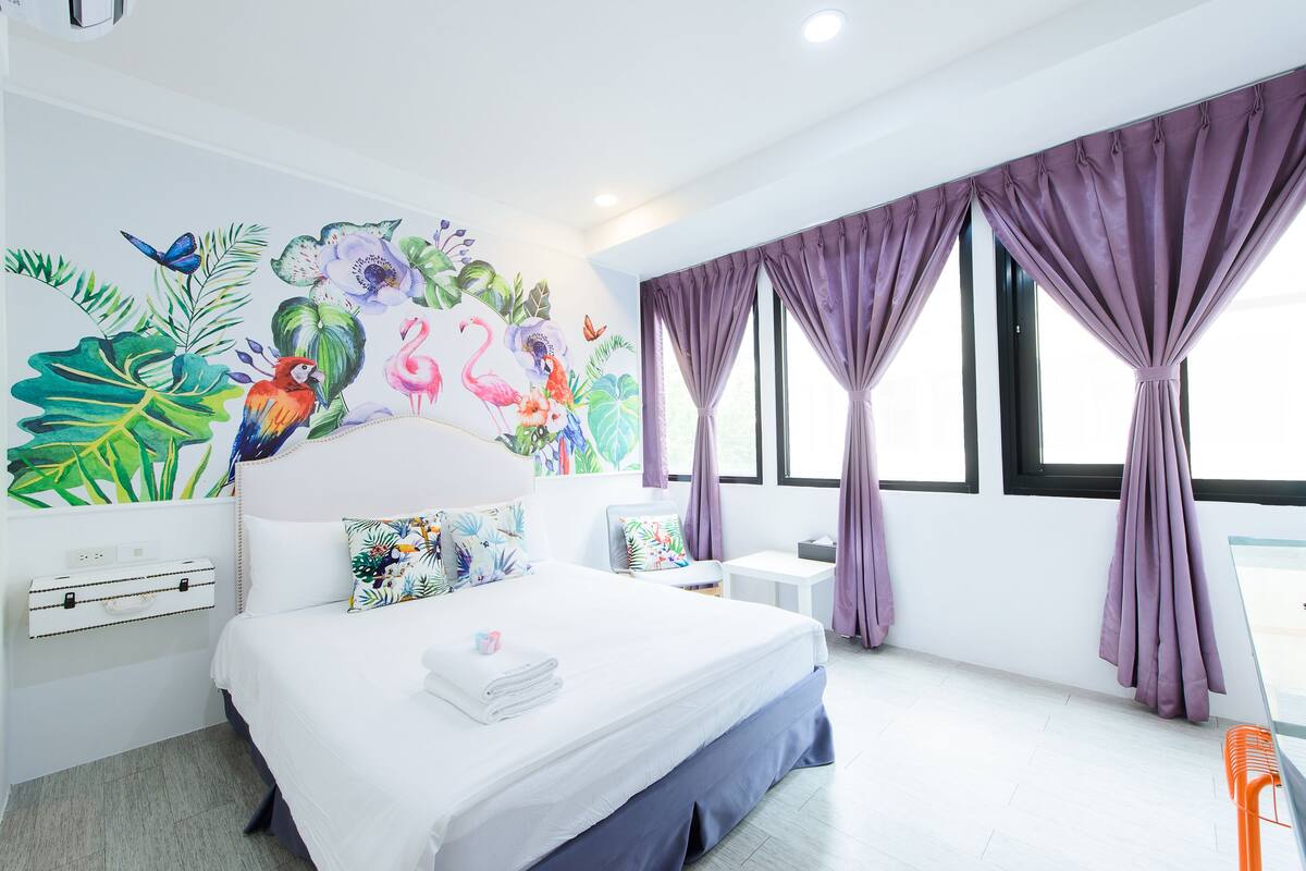 A modern hotel room with a large bed, a colorful mural on the wall, and large windows with purple curtains.