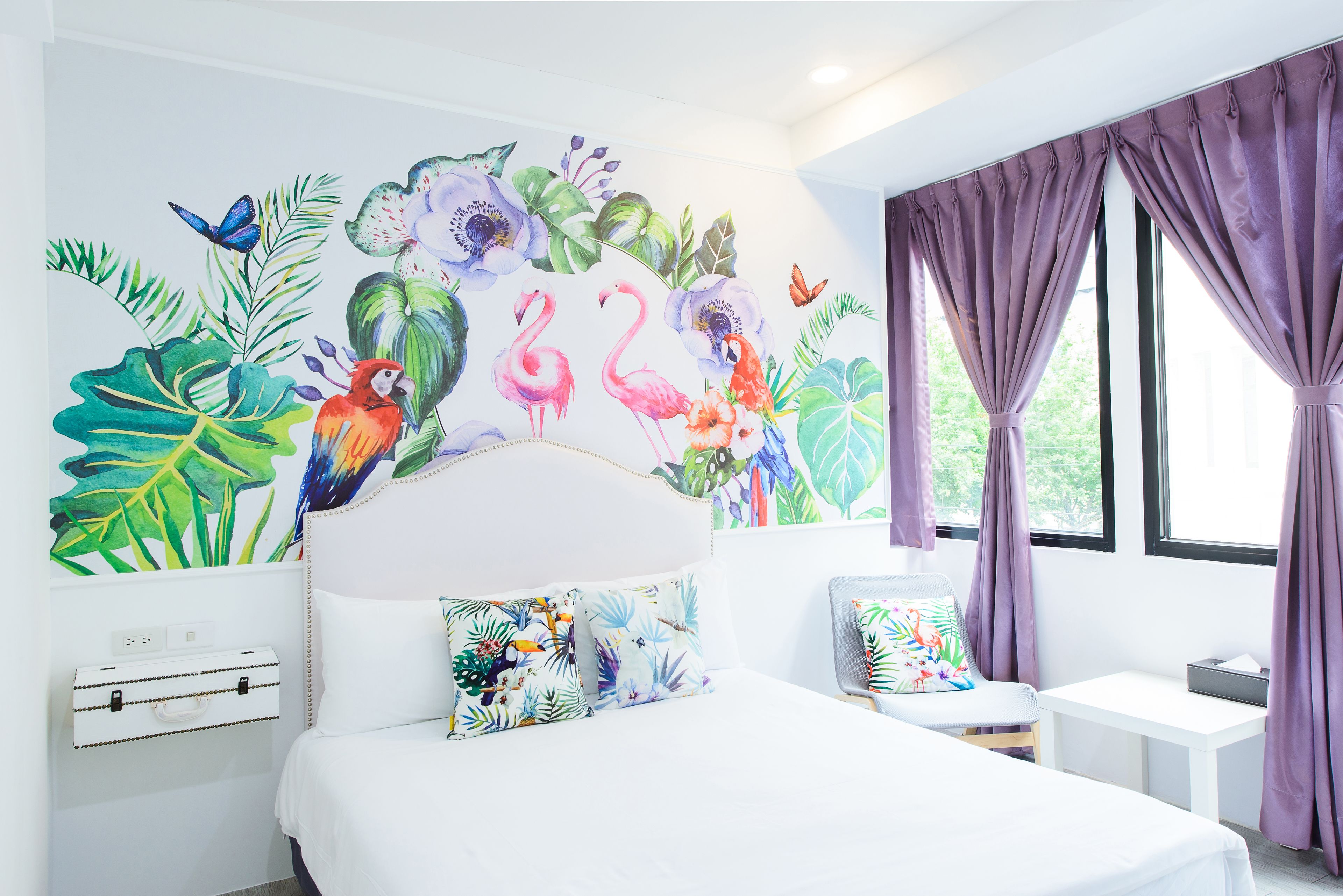 A bedroom with a large bed, a desk, and a chair. There is a window with purple curtains and a wall mural with tropical plants and animals.