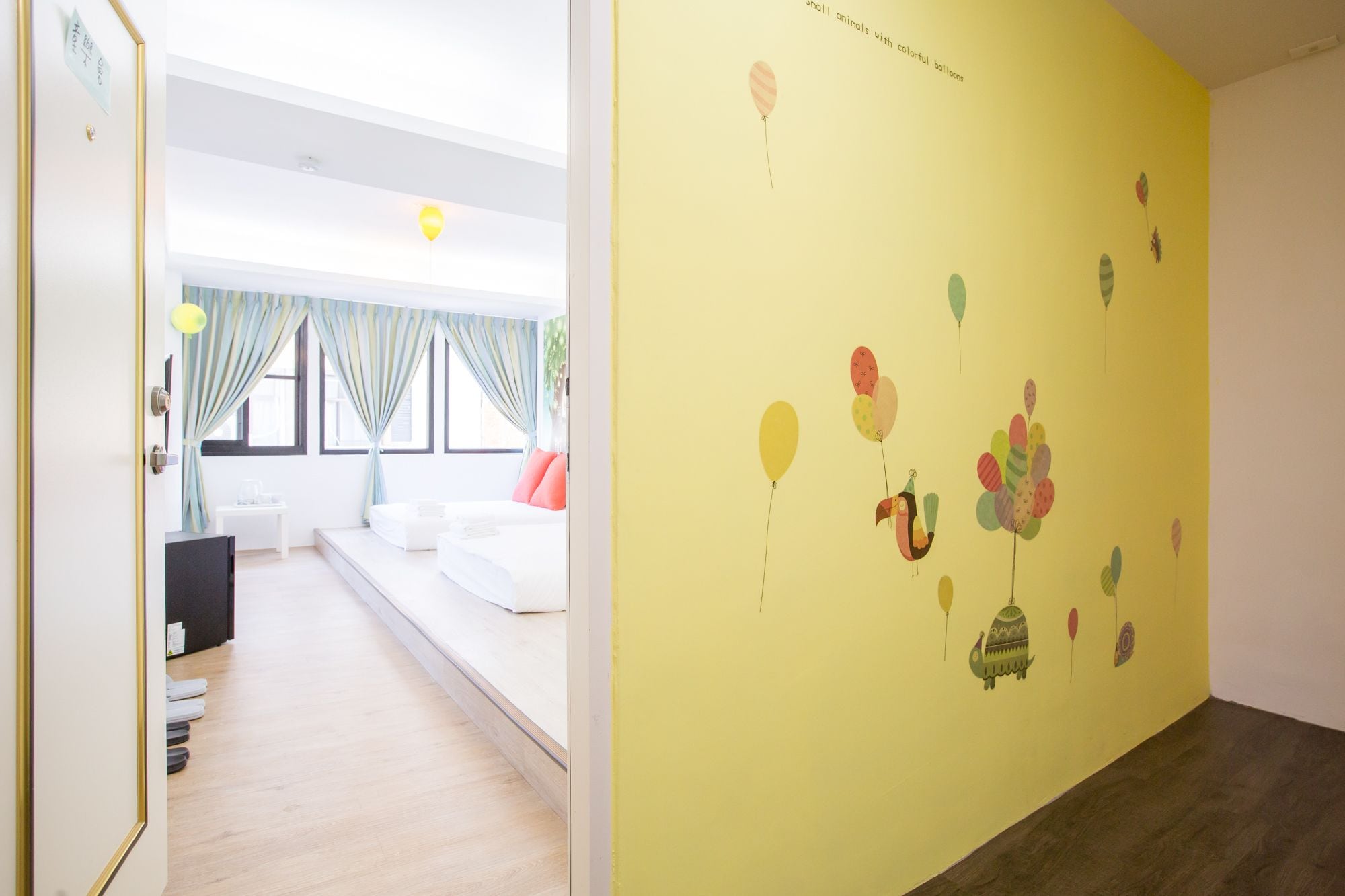 A hotel room with a bed, a desk, and a window with curtains. The room has a yellow wall with a balloon mural.