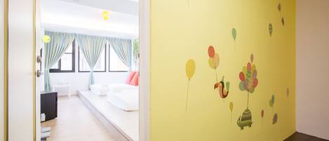 A hotel room with a bed, a desk, and a window with curtains. The room has a yellow wall with a balloon mural.