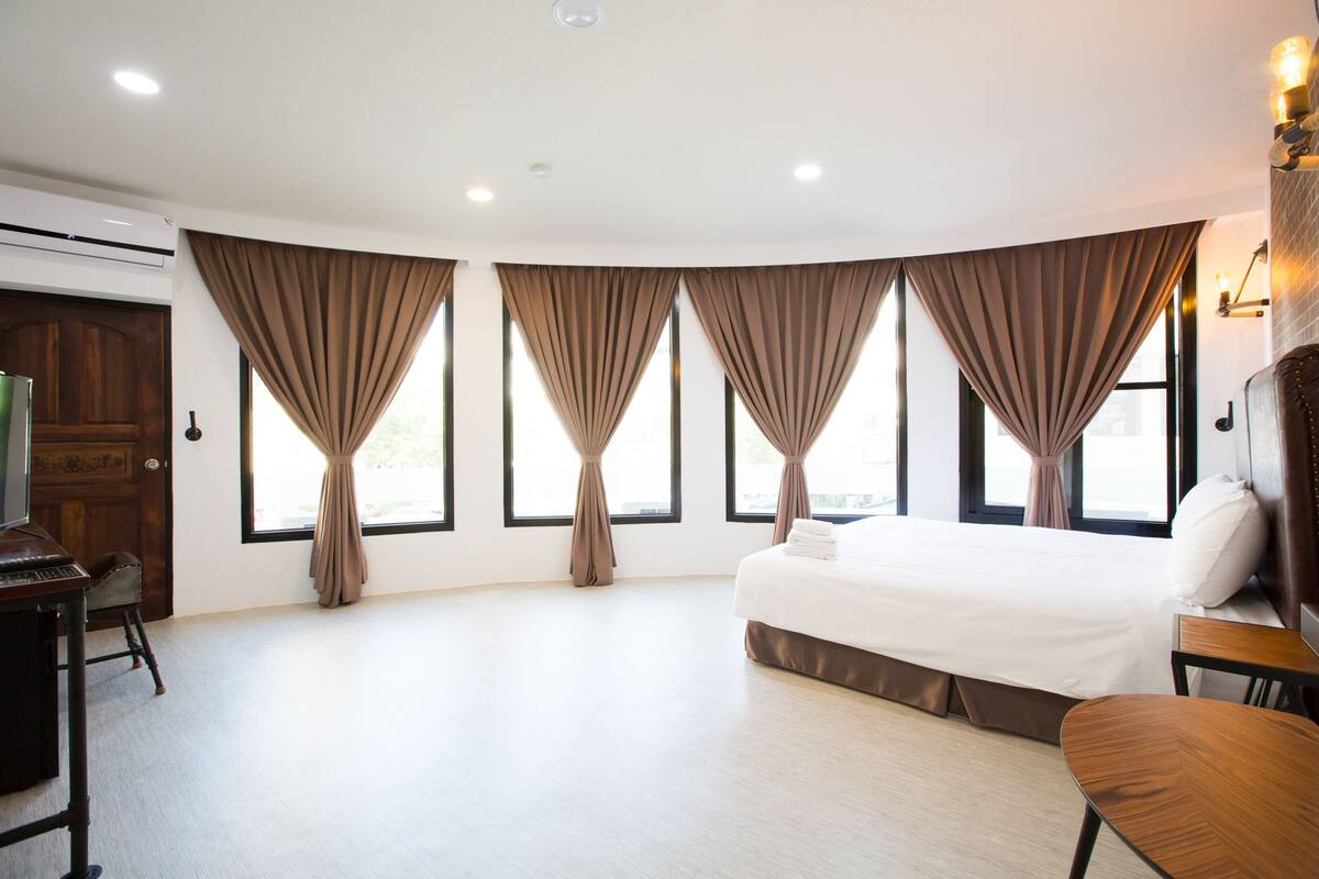 A hotel room with a large bed, a desk, and a chair. There are curtains on the windows, and an air conditioning unit is visible on the wall.