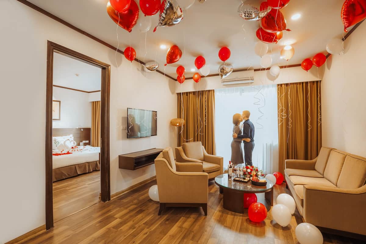 A hotel room with a wooden floor, beige sofas, a coffee table, and a bed with a red and white bedspread.