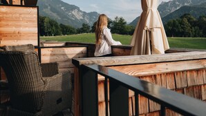 A wooden deck with wicker furniture and a table, overlooking a grassy field and mountains. - Klosterhof - Alpine Hideaway & Spa (Bayerisch Gmain)