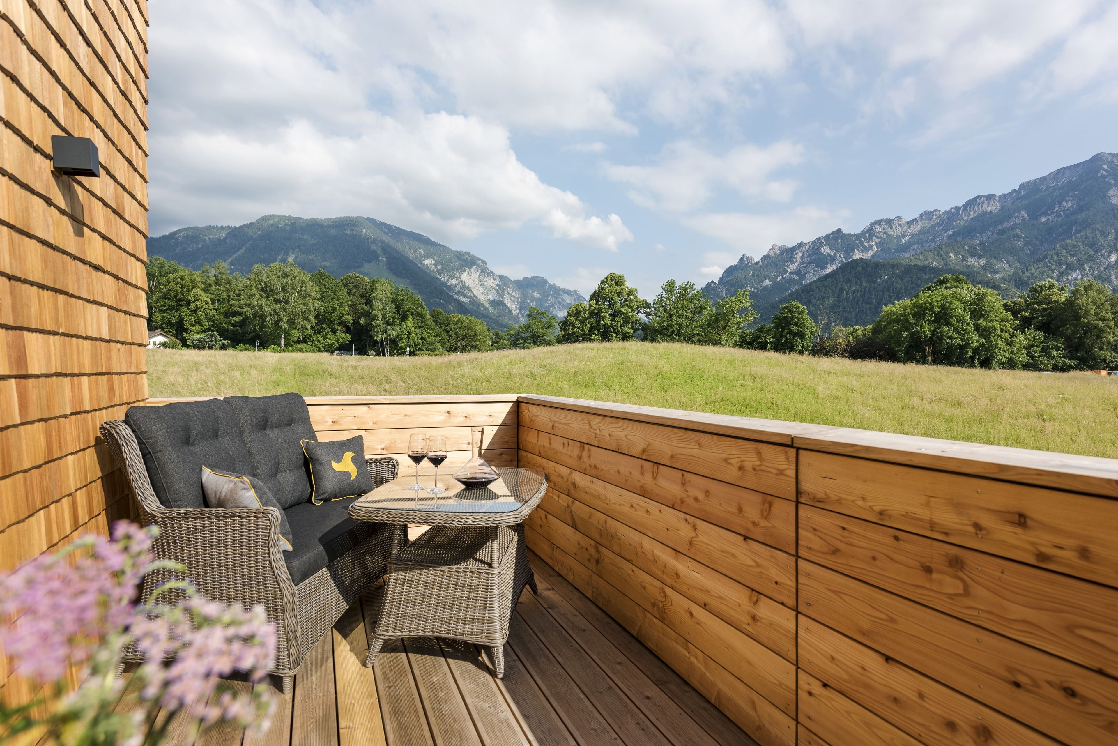 Una terraza de madera con mobiliario de mimbre y una mesa, con vistas a un paisaje montañoso.