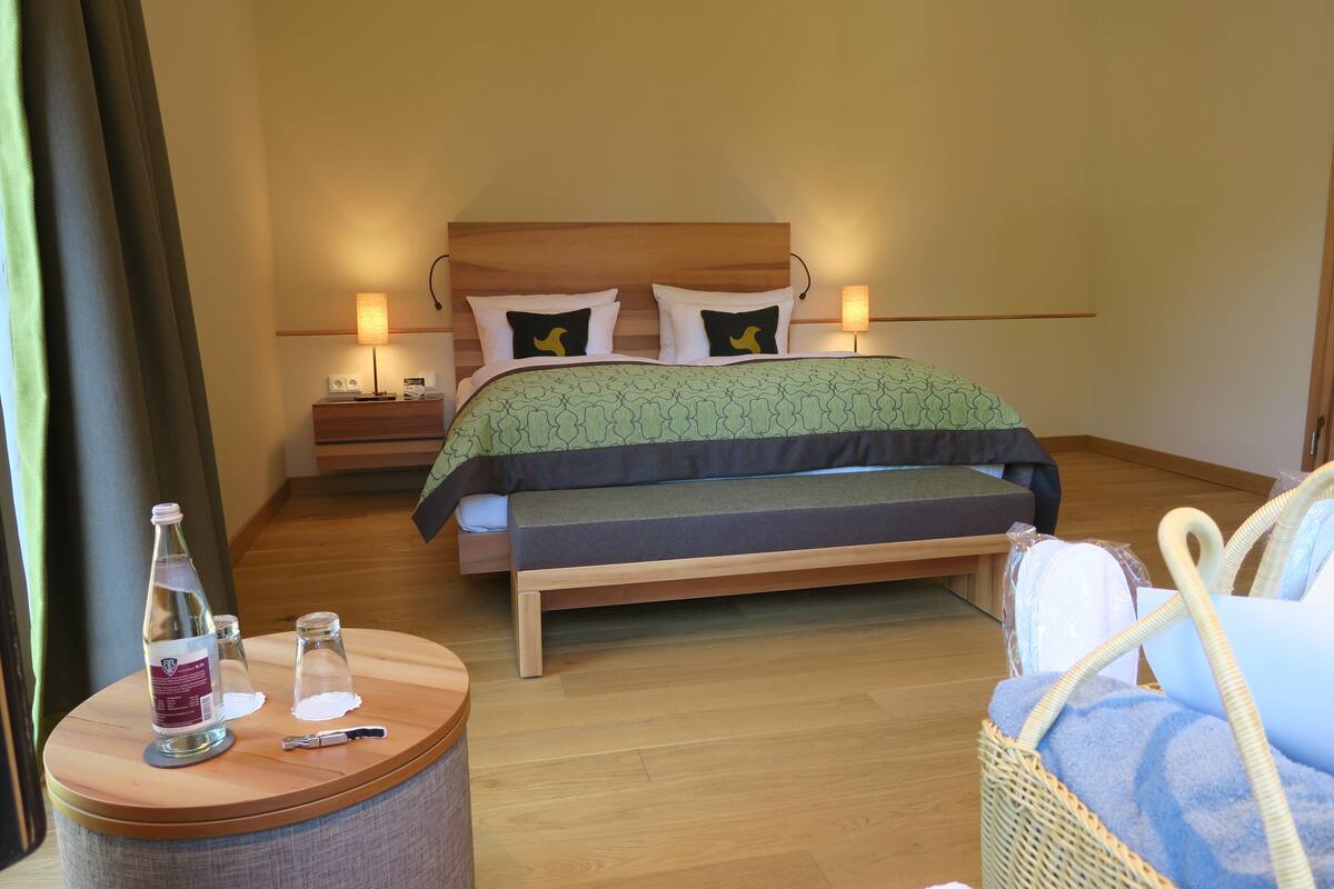 A neatly arranged bedroom with a bed, bedside lamps, a bedside table with a bottle, glasses, and a remote control.