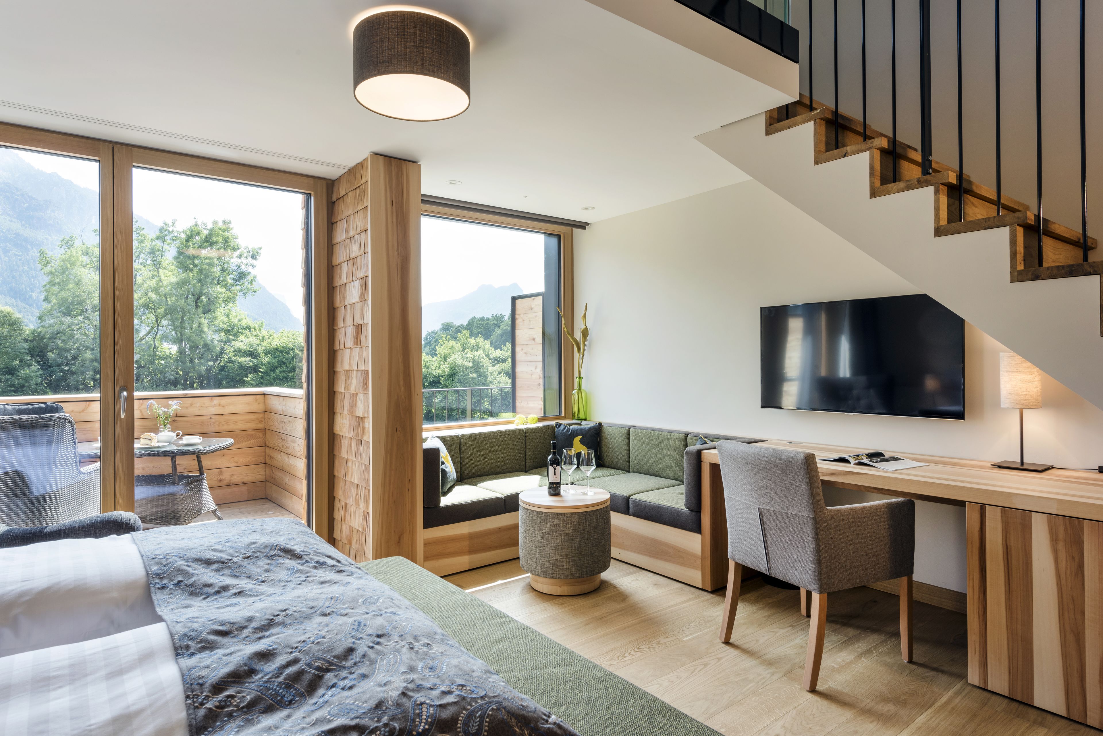 Ein modernes Hotelzimmer mit großem Fenster und Bergblick, einer gemütlichen Sitzecke, einem Flachbildfernseher und einem Holztisch.
