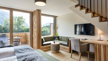 Ein modernes Hotelzimmer mit großem Fenster und Bergblick, einer gemütlichen Sitzecke, einem Flachbildfernseher und einem Holztisch.