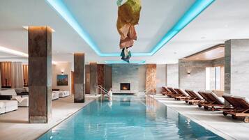 Ein moderner, überdachter Swimmingpool mit klarem, blauem Wasser, Liegestühlen entlang der Seiten und einem Wasserfall im Zentrum.