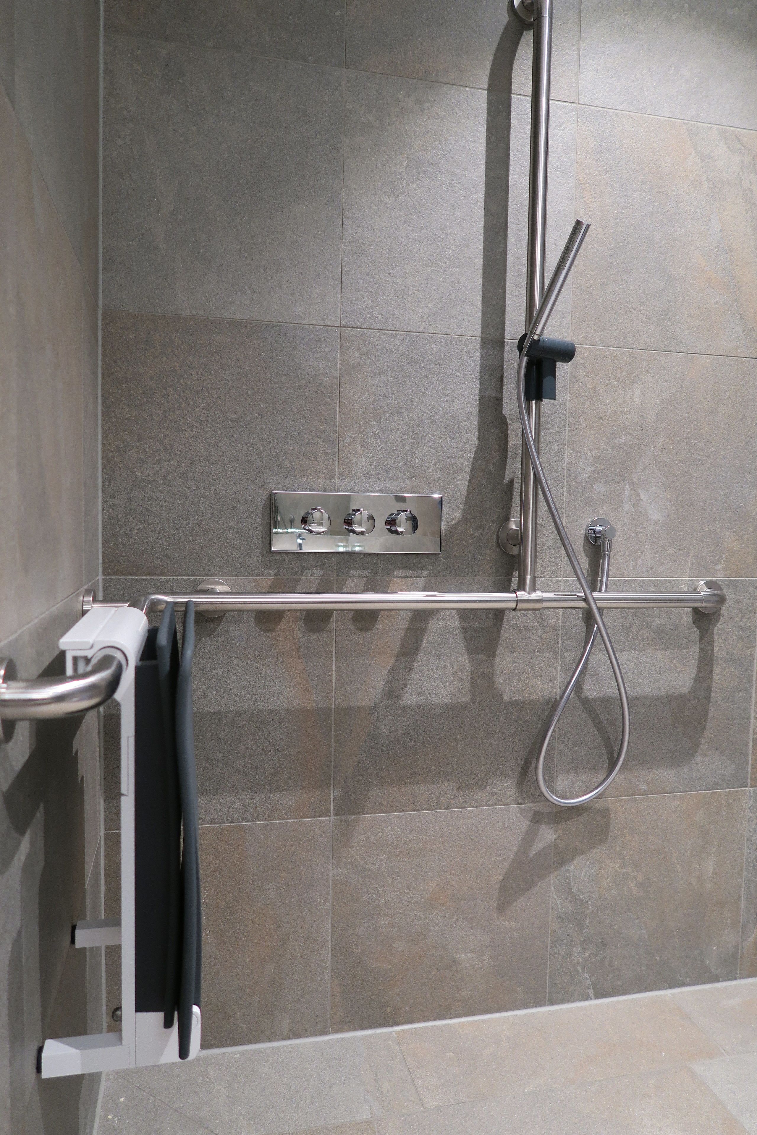 Un área de ducha con cabezal de ducha manual, panel de control en la pared y barra de apoyo.