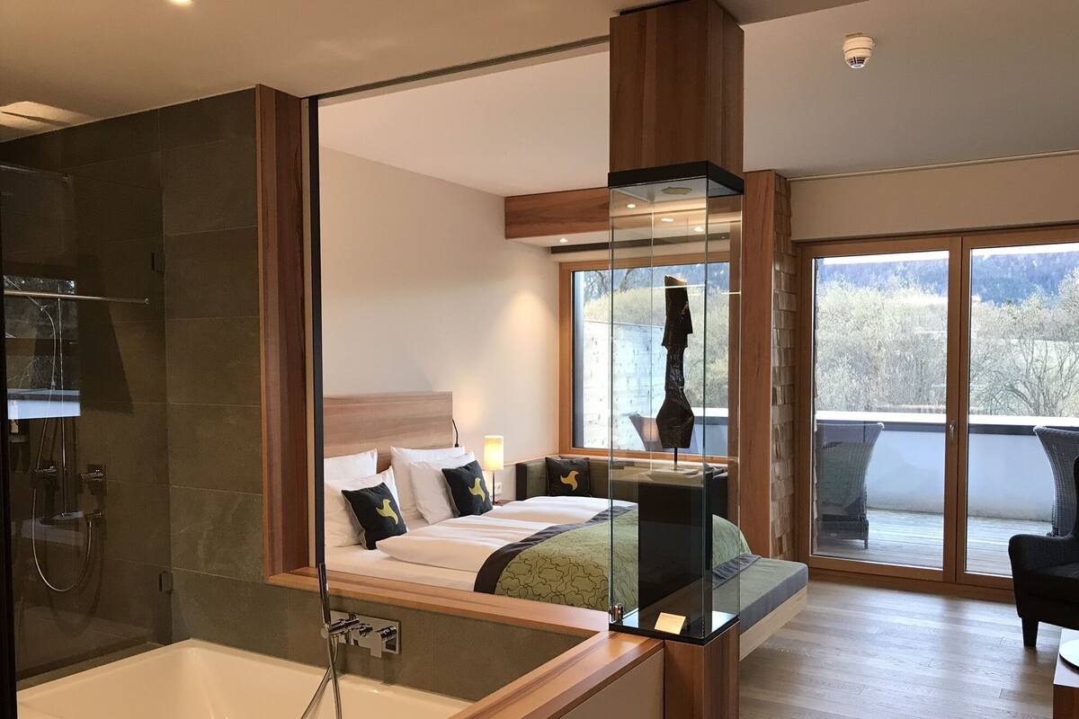 A modern hotel room with a glass-enclosed shower, a bed with a view of the outdoors, and a balcony with wicker furniture.