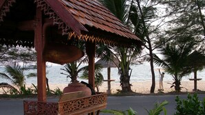 Um gazebo de madeira com esculturas detalhadas, uma palmeira e uma praia ao fundo.