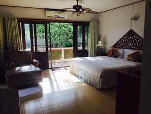 Ein Schlafzimmer mit Bett, Sessel, Deckenventilator, Klimaanlage, Nachttisch und Blick ins Grüne durch das Fenster.
