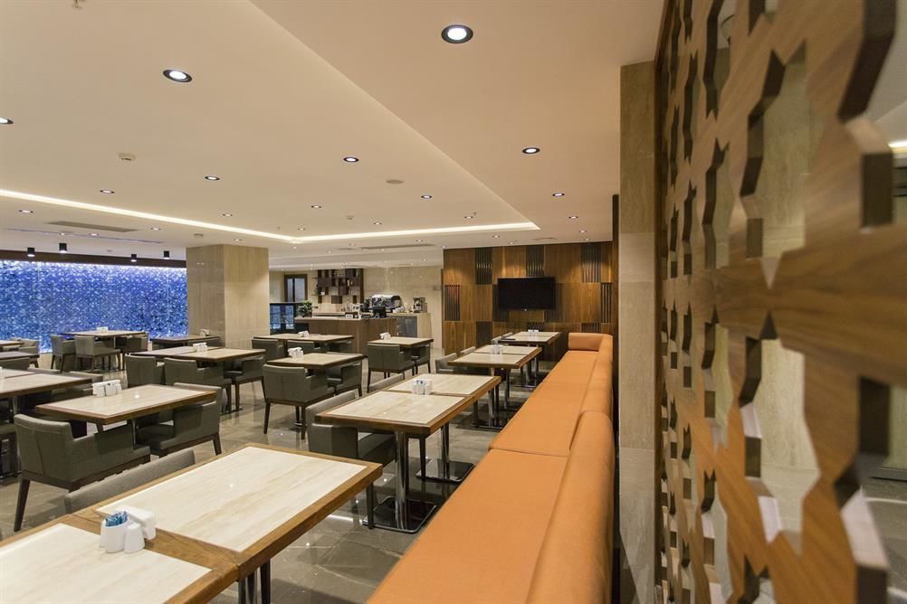 A modern restaurant with a long counter and multiple tables, a wooden accent wall, and a view of the city.