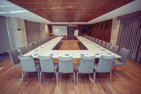 Una sala de conferencias con una larga mesa, sillas grises, una pantalla de proyector y una pizarra.