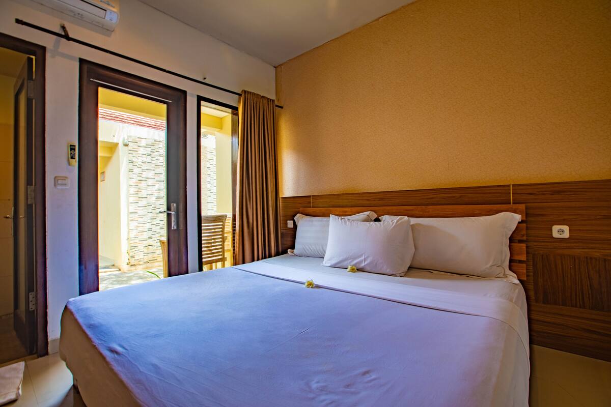 A hotel room with a large bed, wooden headboard, and a view of a courtyard through an open door.