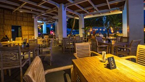 Een openluchtrestaurant met houten tafels en stoelen, een overdekt terras met uitzicht op zee, en sfeerverlichting.