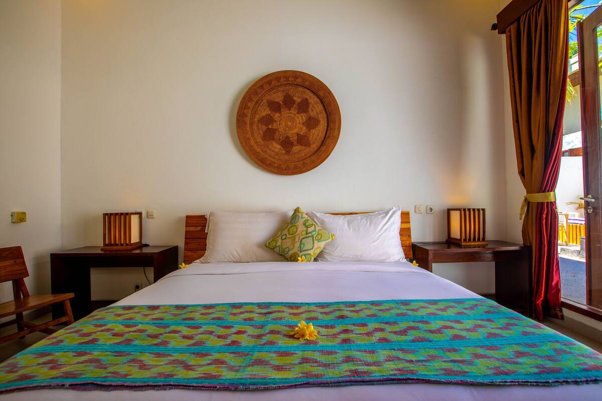 A hotel room with a bed, a wall-mounted decorative plate, a colorful bedspread, and a window with curtains.