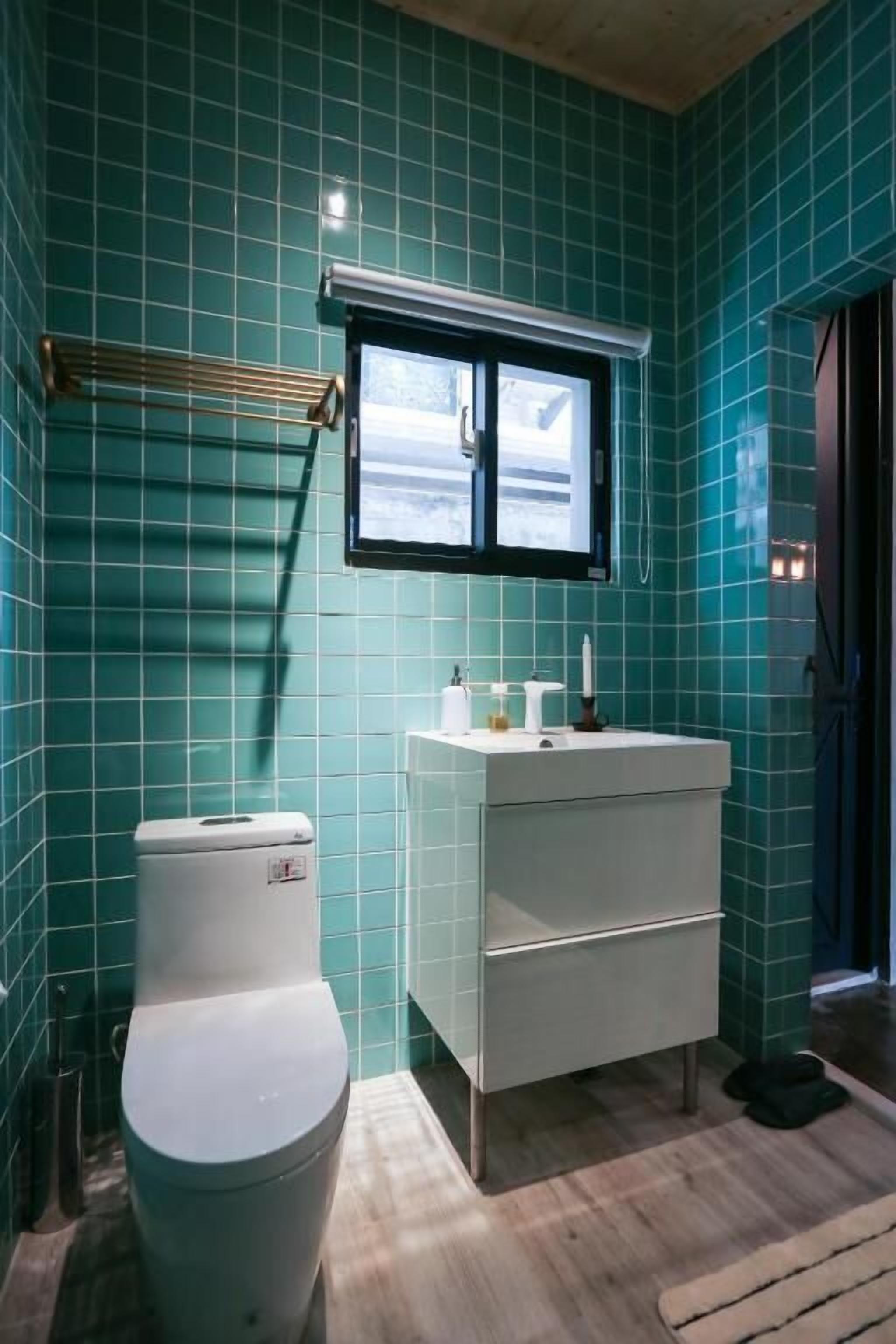 A bathroom with teal tiles, a white toilet, a white sink with a mirror above it, and a wooden floor.
