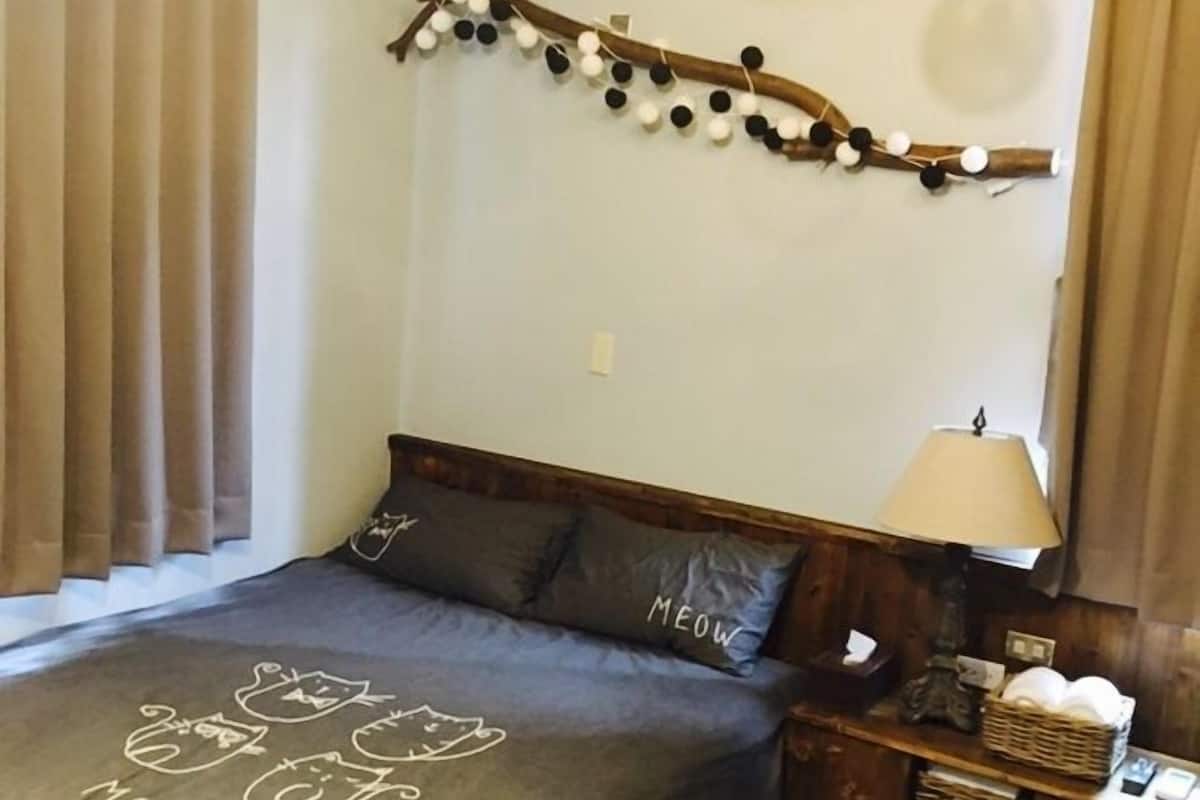 A cozy bedroom with a wooden ceiling, a bed with a grey bedspread featuring cat illustrations, a bedside table with a lamp, and a wall-mounted decoration of black and white balls on a branch.