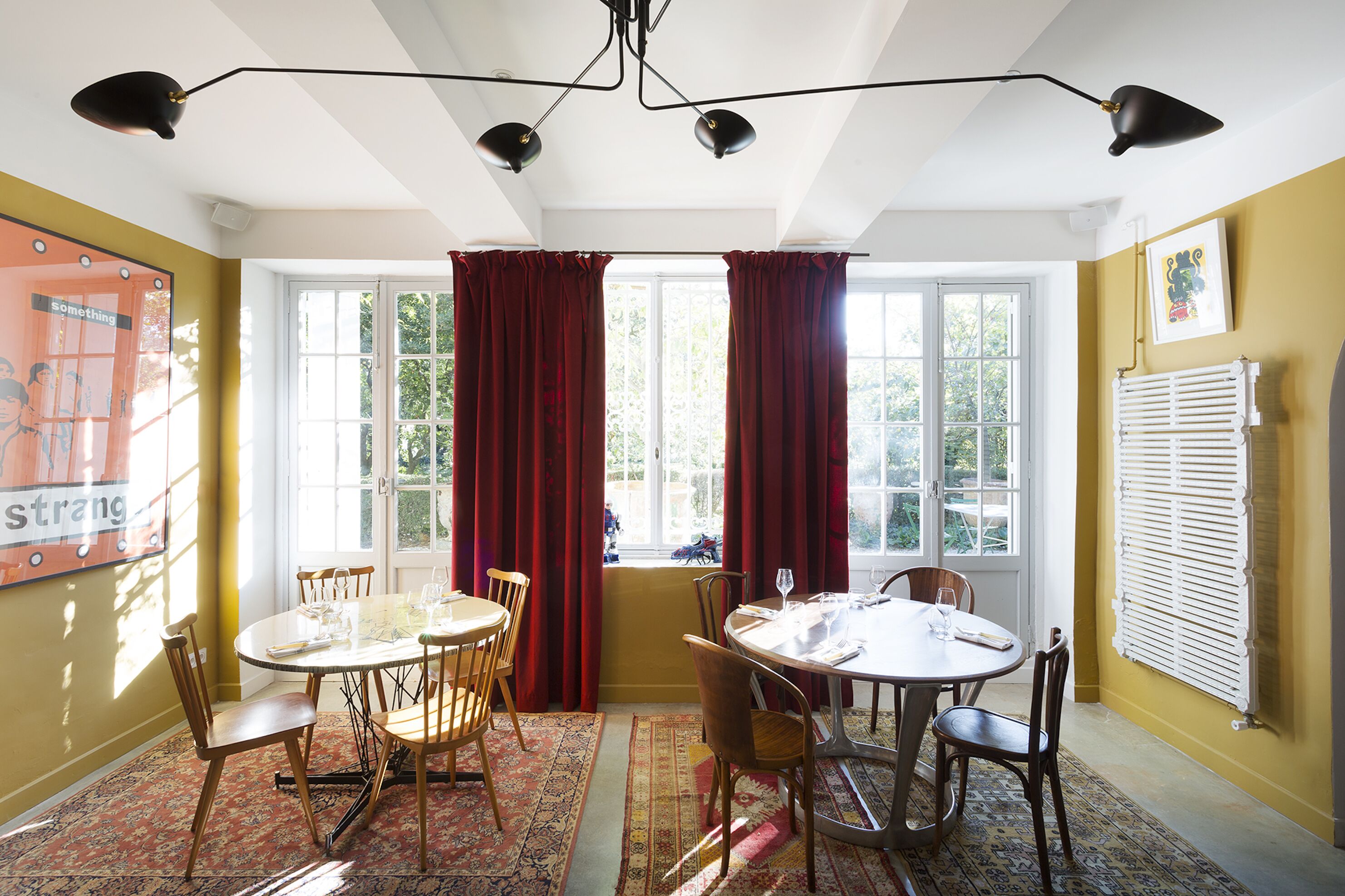 Uma sala de jantar com duas mesas redondas, cortinas vermelhas e vista para árvores através de grandes janelas.