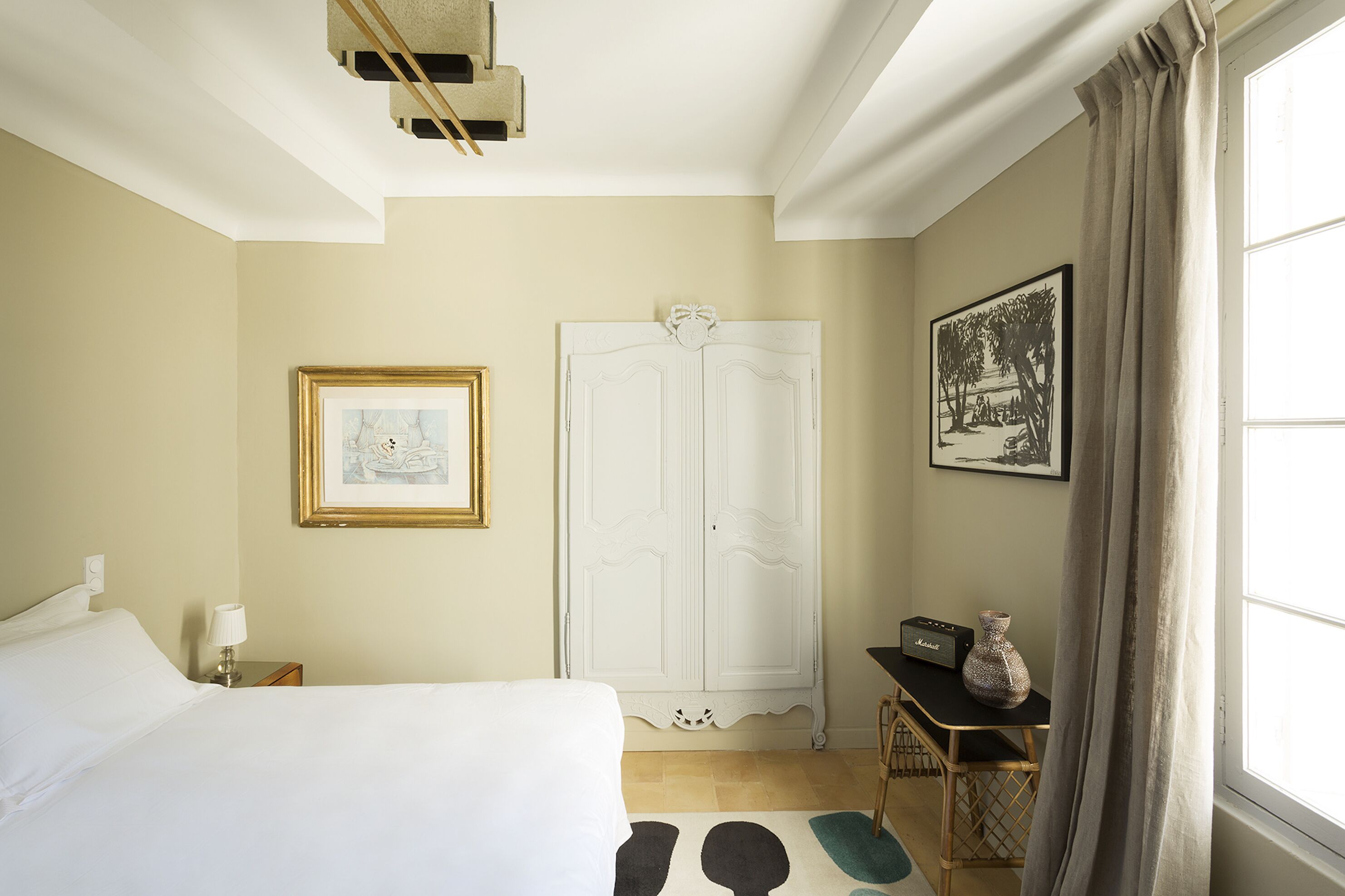 Ein Hotelzimmer mit Bett, Kleiderschrank, einem gerahmten Bild, einem Tisch mit Radio und Vase sowie einem Fenster mit Vorhängen.