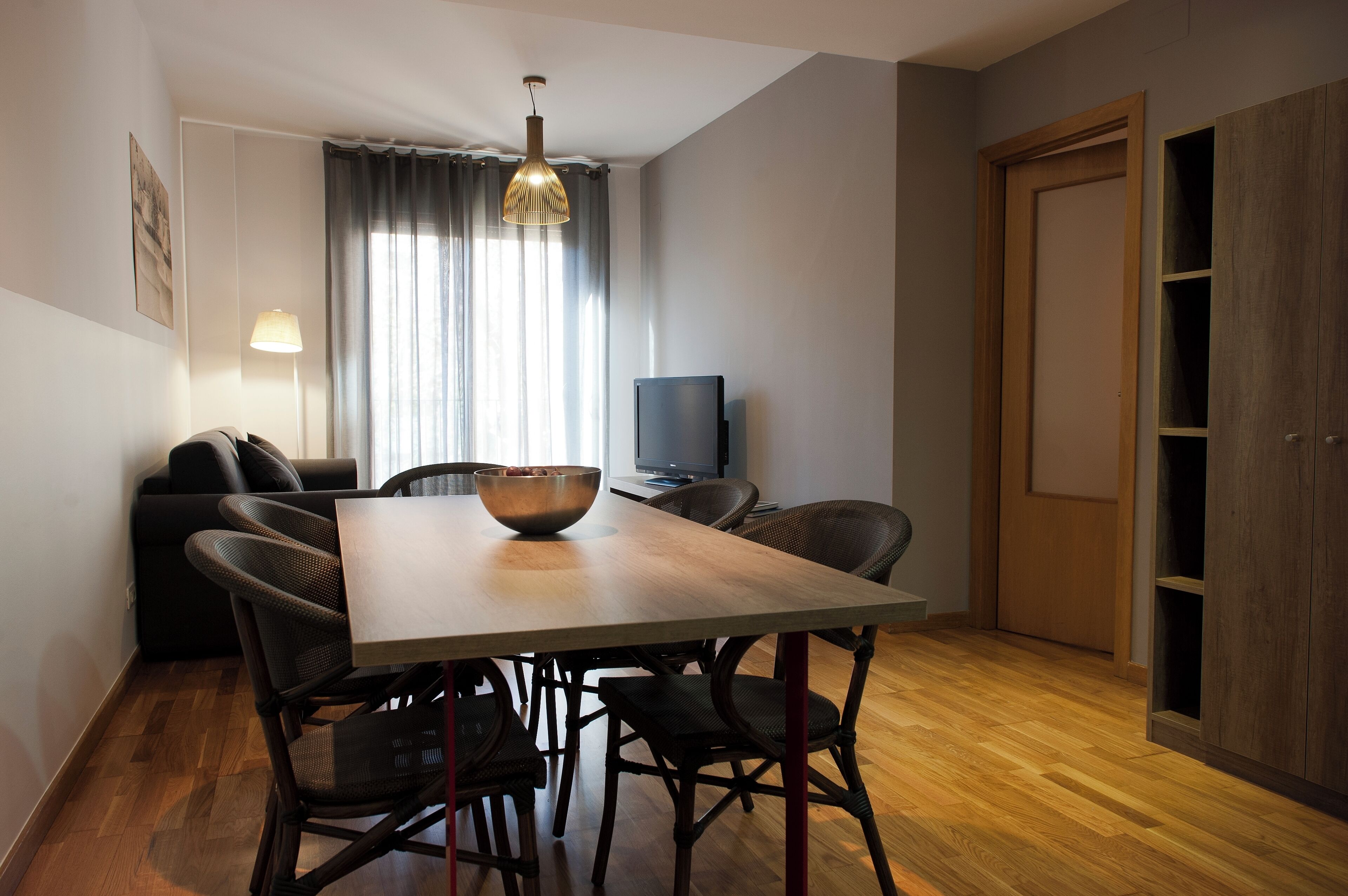 Een eetruimte met een houten tafel, rieten stoelen, een televisie en een groot raam met gordijnen.