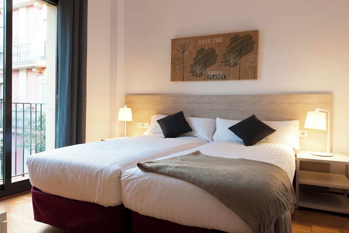 A double bed with white linens, two black pillows, and a green throw blanket. There are two bedside tables with lamps. A wall art piece above the bed reads "SAVE THE GREEN PLANET."