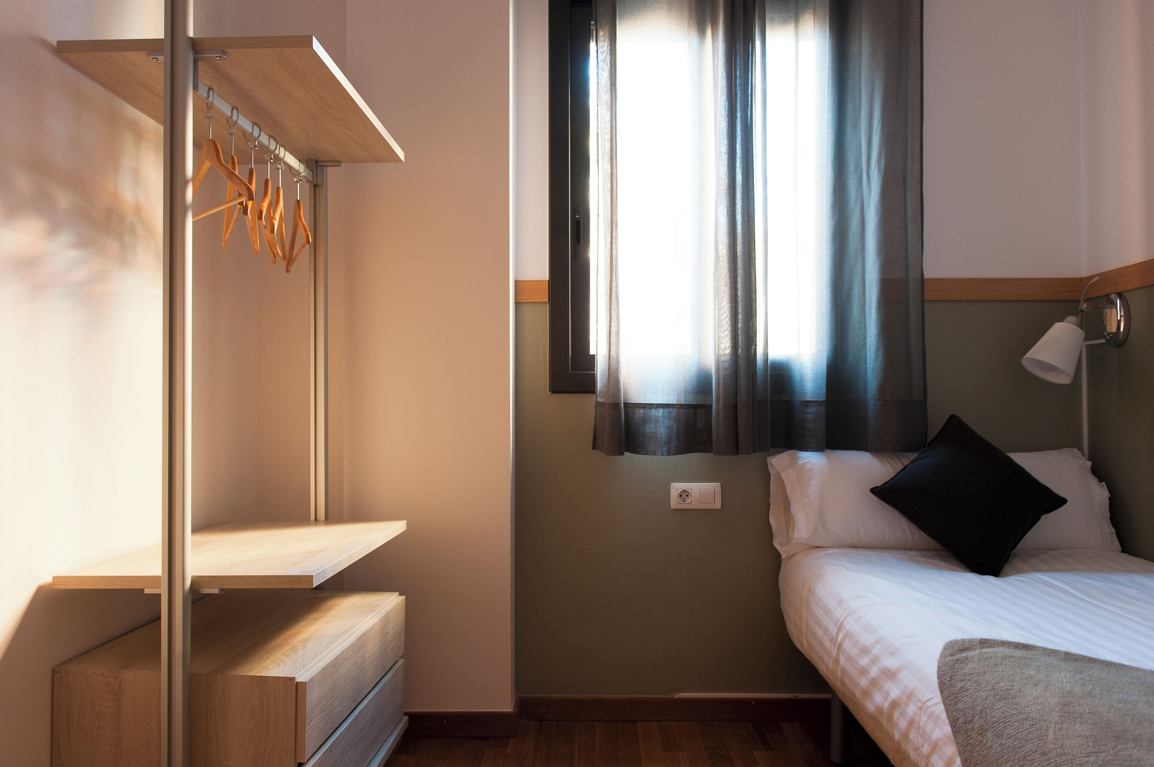 Un dormitorio con cama, mesita de noche, estantería de madera, ventana con cortinas y lámpara de pared.