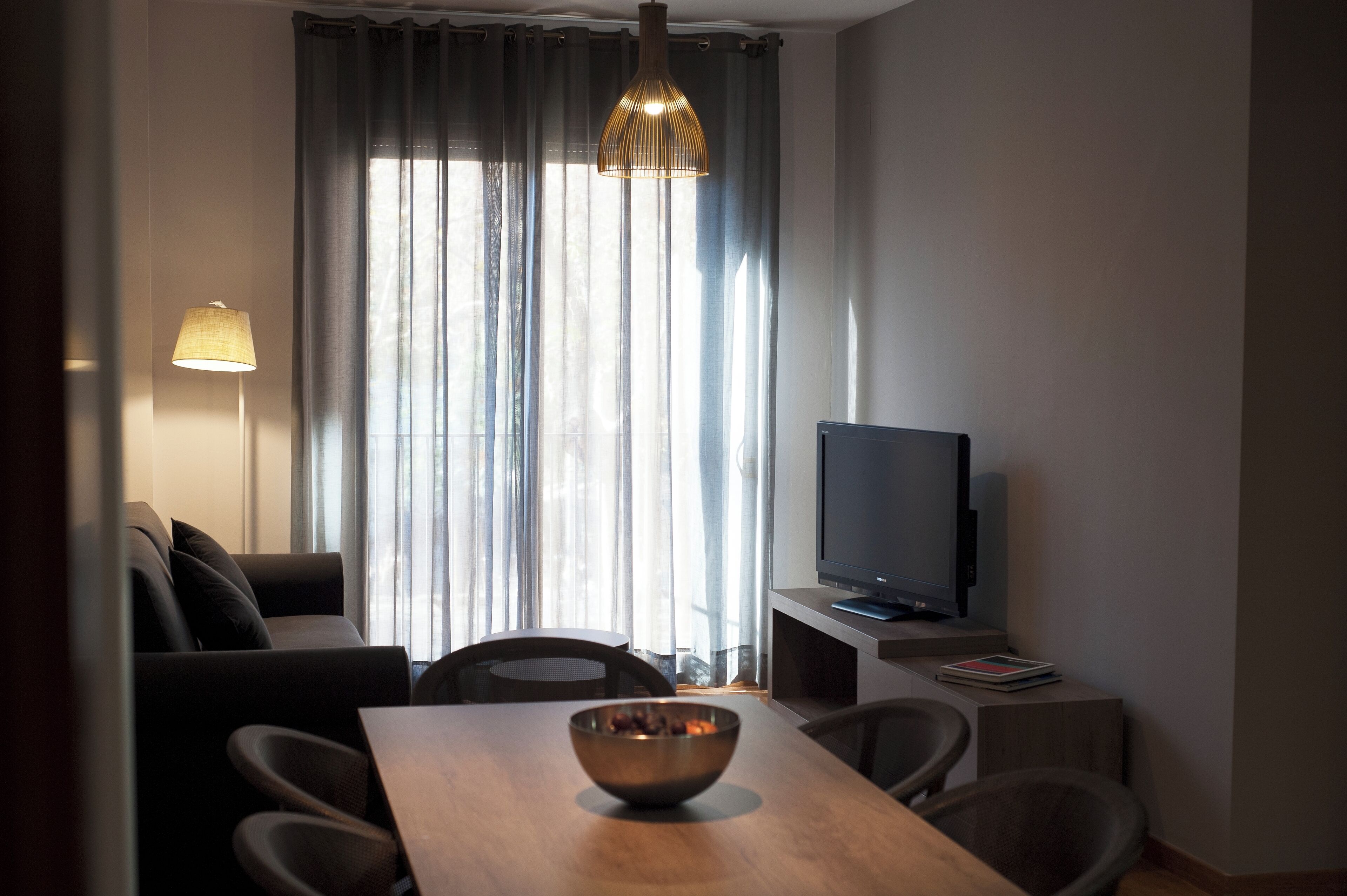 Una sala de estar moderna con un sofá, una mesa de comedor, un televisor sobre un mueble bajo y un amplio ventanal con cortinas.