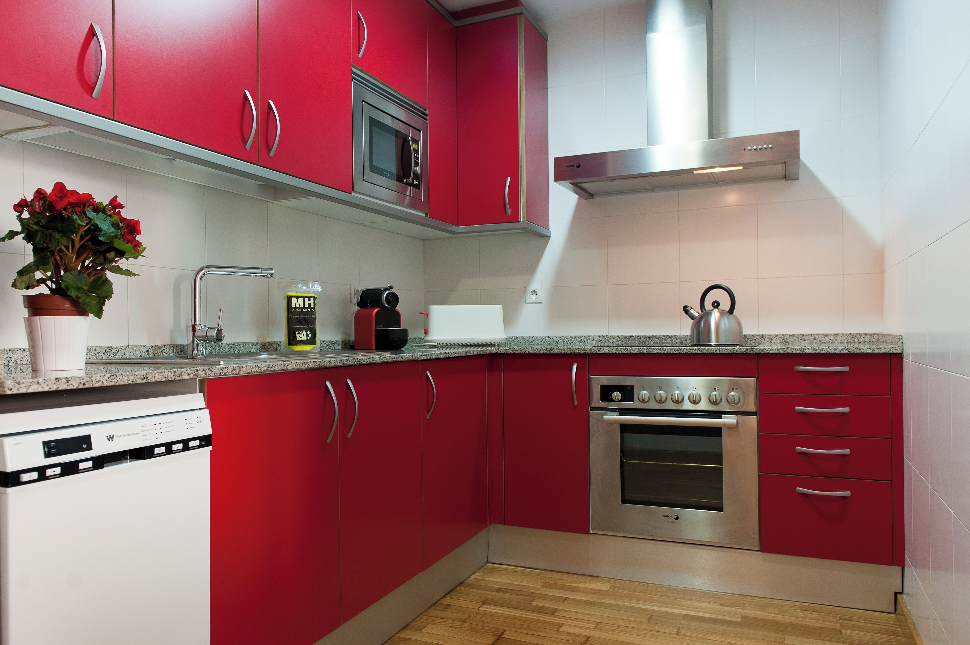 Una cocina con armarios rojos, electrodomésticos de acero inoxidable, un hervidor y una planta en maceta.