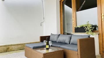 Un canapé en osier avec des coussins, une table basse avec un plateau en verre et une bouteille de vin.