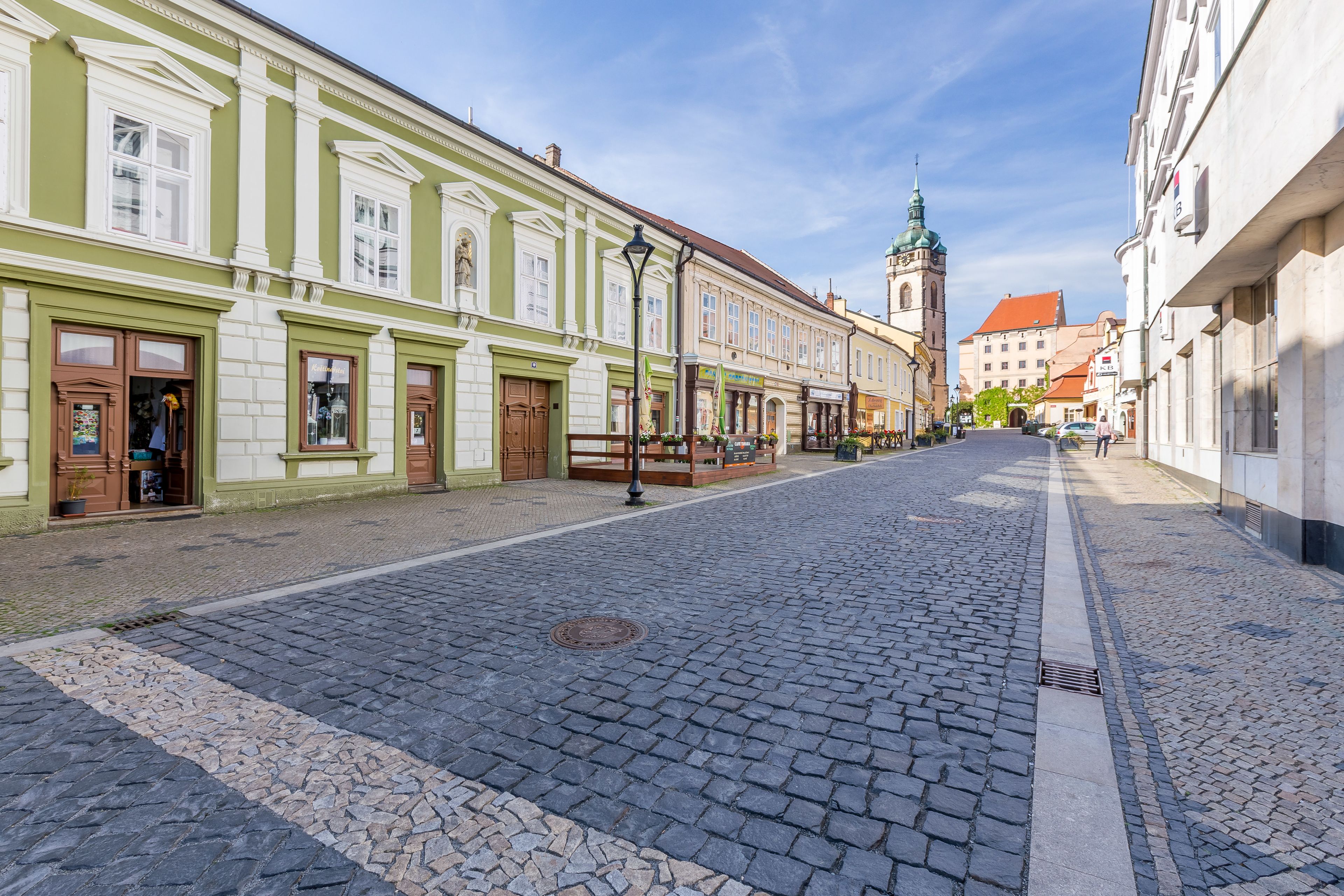 A cobblestone street lined with historic buildings, a church spire in the distance, and a clear blue sky.