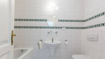 Un baño con lavamanos, inodoro y bañera blancos. Hay una toalla colgada en la pared y un espejo sobre el lavamanos.