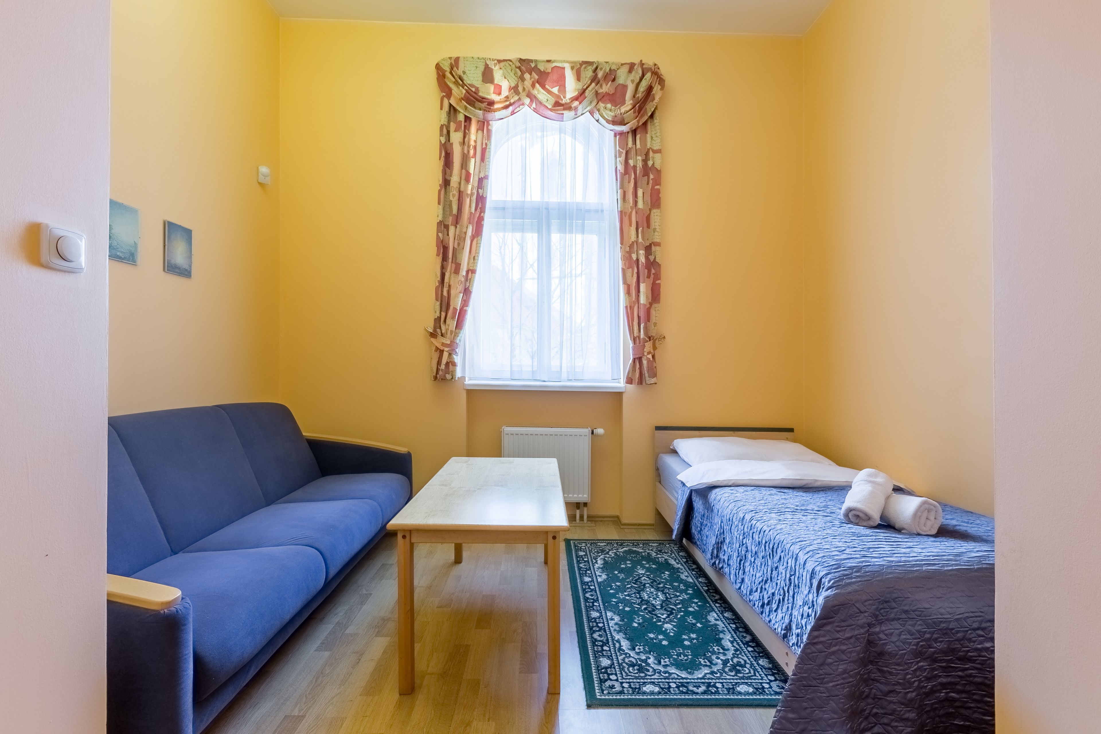 A small, bright room with a sofa, a bed, a table, and a window with curtains.