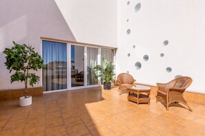 A patio area with a white wall, a potted plant, wicker chairs, a small table, and a sliding glass door leading to an interior space.